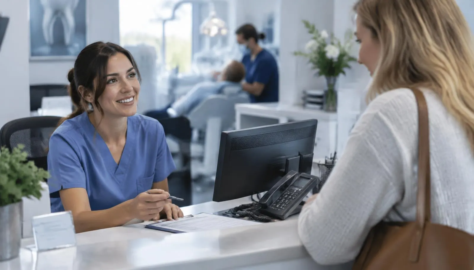 Dental clinic receptionist speaking with a patient at the front desk beside a desk phone and computer